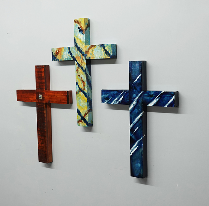 Three handmade wooden crosses — cedar, painted, and blue — displayed on a white wall